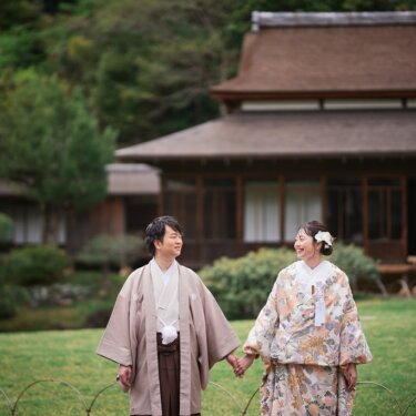三渓園の茅葺屋根の大きな建物を背景に、芝生の上で手をつなぎ見つめ合う色打掛の花嫁と羽織袴の新郎の和装前撮り