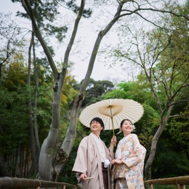 白い和傘を差して木々を見上げ、笑い合う色打掛の花嫁と羽織袴の新郎。庭園の木立で撮る和装フォトウェディング