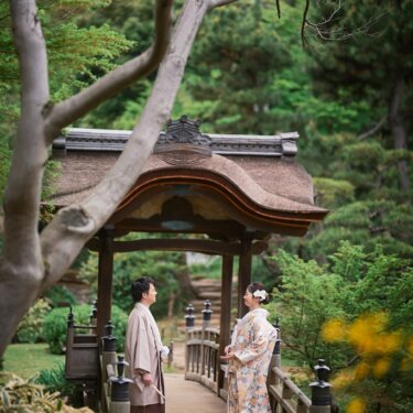 三渓園の唐破風の東屋を背景に、橋の上で見つめ合う新郎新婦。手前の黄色い花がアクセントの和装前撮り