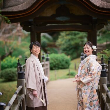 東屋へ続く橋の上で振り返り、カメラに笑顔を向ける羽織袴の新郎と色打掛の花嫁の結婚写真