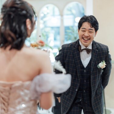 花嫁の背中越しに大笑いするチェックのタキシードの新郎。スタジオで撮るファーストミートの前撮り