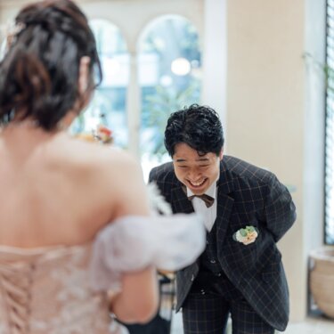 ファーストミートの瞬間、花嫁の背中越しに身をかがめて大笑いする新郎。編み上げのカラードレスのバックスタイルが写る前撮り