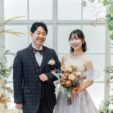 大きなアーチ窓とドライフラワーの装飾を背景に、カメラ目線で笑う新郎新婦。スタジオでのフォトウェディング