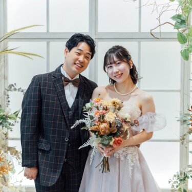 ドライフラワーの装飾に囲まれた窓辺で並ぶ、チェックのスーツの新郎とピンクのカラードレスの花嫁のスタジオフォト