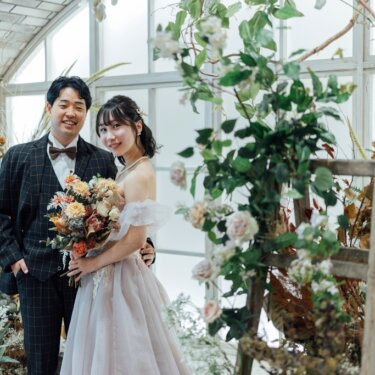 はしごに絡む蔓と花々に囲まれたスタジオセットで、寄り添って笑うチェックのタキシードの新郎とカラードレスの花嫁。洋装前撮り