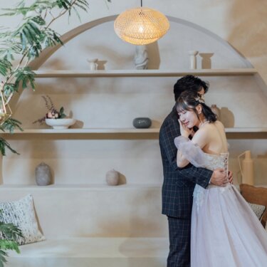ランタン照明が灯るアーチ状の飾り棚の前で、頬を寄せて抱き合う新郎新婦。オフショルダーのカラードレスで撮るスタジオフォト
