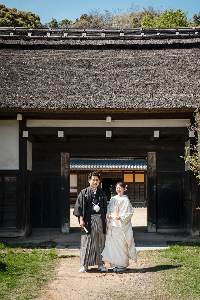 旧横溝家住宅の茅葺屋根の門の前に立つ白無垢と黒紋付袴の新郎新婦
