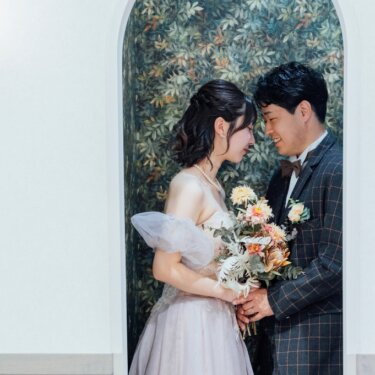 植物柄の壁画とアーチ型の枠を背景に、おでこを寄せ合う新郎新婦。秋色のブーケが映えるスタジオフォト
