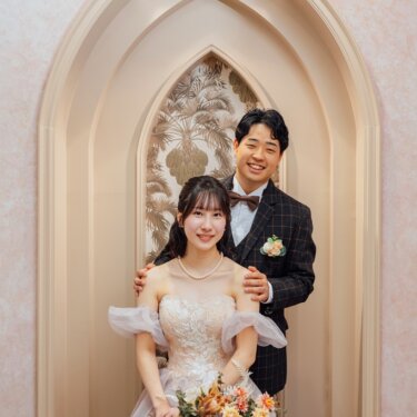 ゴシック調のアーチ型ニッチを背景に、新郎が花嫁の肩に手を添えて笑う一枚。オフショルダーのカラードレスで撮るスタジオフォト