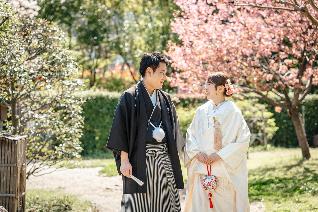 満開の桜の木の下で向き合って微笑む和装の新郎新婦