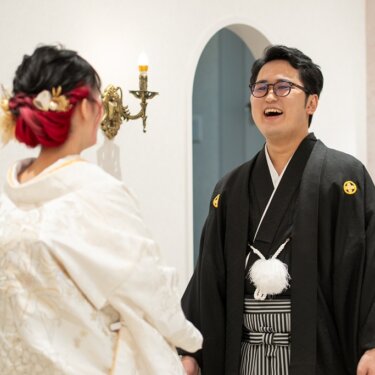 白無垢の花嫁の背中越しに大笑いする紋付袴の新郎。赤い髪色と髪飾りのヘアアレンジが映える、スタジオでのファーストミート前撮り