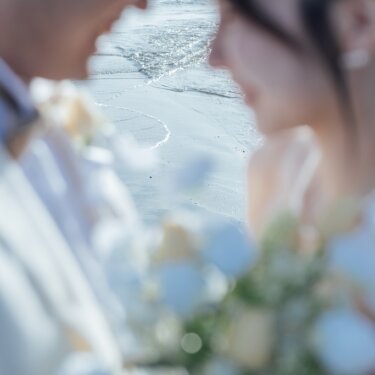 淡いブルーのブーケを前ボケにして、見つめ合う新郎新婦の横顔を写した一枚。波打ち際の光がやわらかな海辺の前撮り