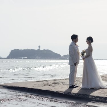 江の島を背景に、砂州の先で向かい合う白いタキシードの新郎とウェディングドレスの花嫁。湘南らしい景色の中で撮る前撮り
