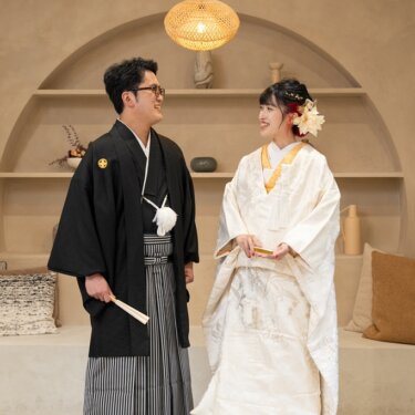 ランタン照明のアーチ棚を背景に、扇を手に見つめ合う紋付袴の新郎と白無垢の花嫁。和洋折衷のスタジオフォト