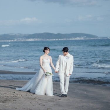 山並みと青い海を背景に、砂浜を手をつないで歩く新郎新婦。サーファーの見える湘南の海辺で撮るフォトウェディング