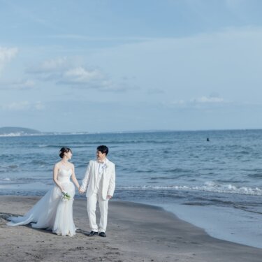 青い空と海を背景に、手をつないで見つめ合う白いタキシードの新郎とウェディングドレスの花嫁。七里ヶ浜で撮る開放的な前撮り