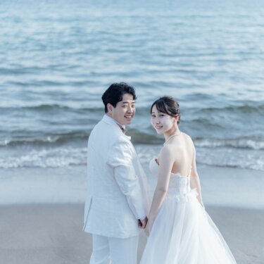 海を背景に振り返って笑う花嫁と新郎。背中が大きく開いたウェディングドレスのバックスタイルが美しい海辺の前撮り