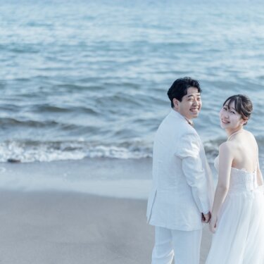 波打ち際で手をつなぎ、振り返って笑う白いタキシードの新郎とウェディングドレスの花嫁。海辺の前撮りの振り返りポーズ