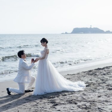 江の島を望む波打ち際で片膝をつき、花嫁に指輪を差し出してプロポーズを再現する新郎。湘南の海辺のフォトウェディング