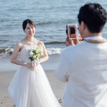 白いタキシードの新郎がカメラで、ブーケを持つウェディングドレスの花嫁を撮影する一枚。海辺の前撮りのほほえましいシーン
