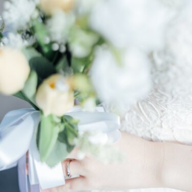 指輪をつけた手で持つ白とクリーム色のブーケのクローズアップ。リボンとレースのドレスが写る前撮りの手元カット