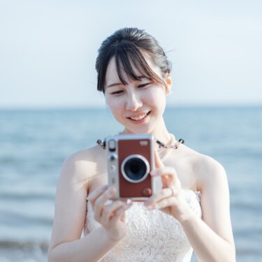 波打ち際でカメラを構えて微笑むウェディングドレスの花嫁。レトロなカメラを小物にした海辺の前撮り