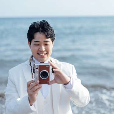 海を背景に、首から下げたカメラを構えて笑う白いタキシードの新郎。撮り合いっこの演出で撮る海辺のフォトウェディング