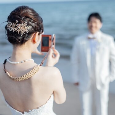 花嫁の後ろ姿越しに、カメラで新郎を撮る構図。ビジューの髪飾りと編み込みのヘアアレンジが映える海辺の前撮り