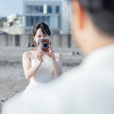 新郎の肩越しに、インスタントカメラを構えるウェディングドレスの花嫁を写した一枚。海辺で撮り合うナチュラルな前撮り