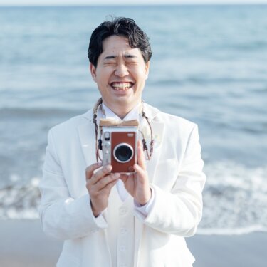海を背景に、インスタントカメラを構えて満面の笑みを浮かべる白いタキシードの新郎。小物を使った遊び心のある前撮り
