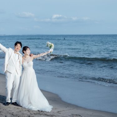 青い海を背景に、ブーケを掲げて両腕を大きく広げる新郎新婦。はしゃぐ姿が楽しい海辺の前撮り
