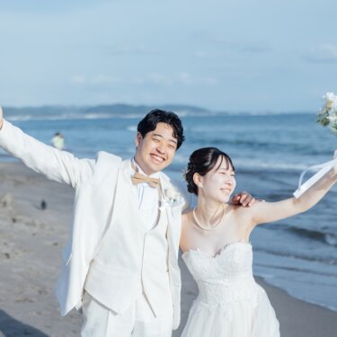 海水浴客の見える夏の砂浜で、ブーケを掲げて手を振る白いタキシードの新郎と花嫁。七里ヶ浜の開放感あふれるフォトウェディング