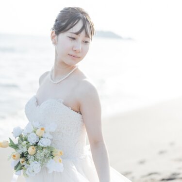 江の島を遠くに望む砂浜で、パールネックレスと黄色のブーケが映えるウェディングドレスの花嫁のソロショット。明るい光の海辺の前撮り
