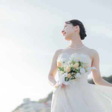 ウェディングドレスの裾をつまみ、空を見上げて微笑む花嫁のソロショット。パールのネックレスが映える海辺の前撮り