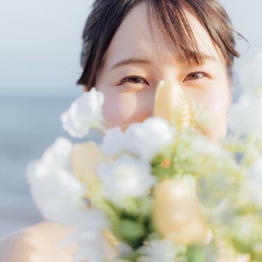 白とクリーム色のブーケで顔の下半分を隠し、目元で笑う花嫁のクローズアップ。海辺の前撮りで人気の顔隠しポーズ