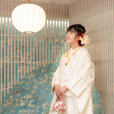 丸い和紙の照明と水色の花柄の壁を背景に、手まりを持って遠くを見る白無垢の花嫁のソロショット。赤い髪飾りのヘアアレンジが映える前撮り