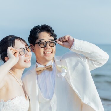 海辺でお揃いの黒縁メガネに手を添えて笑う、白いタキシードの新郎とウェディングドレスの花嫁。遊び心のある前撮りポーズ