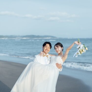 青い海を背景に、ブーケを掲げた花嫁をお姫様抱っこする白いタキシードの新郎。笑顔がはじける海辺の前撮り