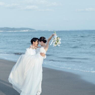 新郎が花嫁をお姫様抱っこし、ブーケを掲げて笑い合う一枚。青い海を背景にした湘南のビーチフォトウェディング
