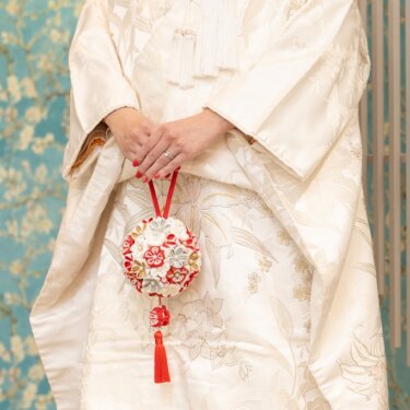 白無垢の花嫁が両手で持つ、赤と白の手まりの小物のクローズアップ。花柄の壁が映える和装前撮りの手元カット