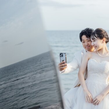海を背景にスマホで自撮りをする、白いタキシードの新郎とウェディングドレスの花嫁。七里ヶ浜で撮るナチュラルな前撮り