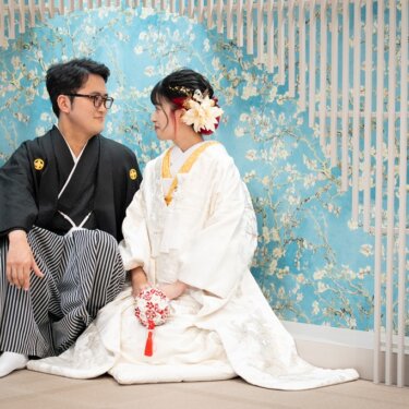 水色の花柄の壁と白い格子を背景に、床に座って見つめ合う新郎新婦。手まりを持つ白無垢の花嫁の和装スタジオ前撮り
