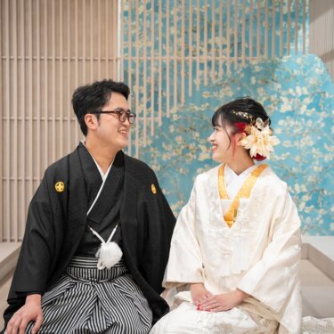 レンガの壁と水色の花柄の格子を背景に、ベンチに座って見つめ合う紋付袴の新郎と白無垢の花嫁。和モダンなスタジオ前撮り