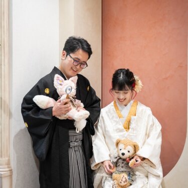 テラコッタ色のアーチ壁を背景に、ぬいぐるみを手に笑う紋付袴の新郎と白無垢の花嫁。ほほえましい和装スタジオフォト