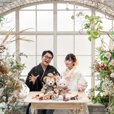 アーチ窓と花々のスタジオでテーブルにぬいぐるみを並べて笑う、黒紋付袴の新郎と白無垢の花嫁のスタジオフォト