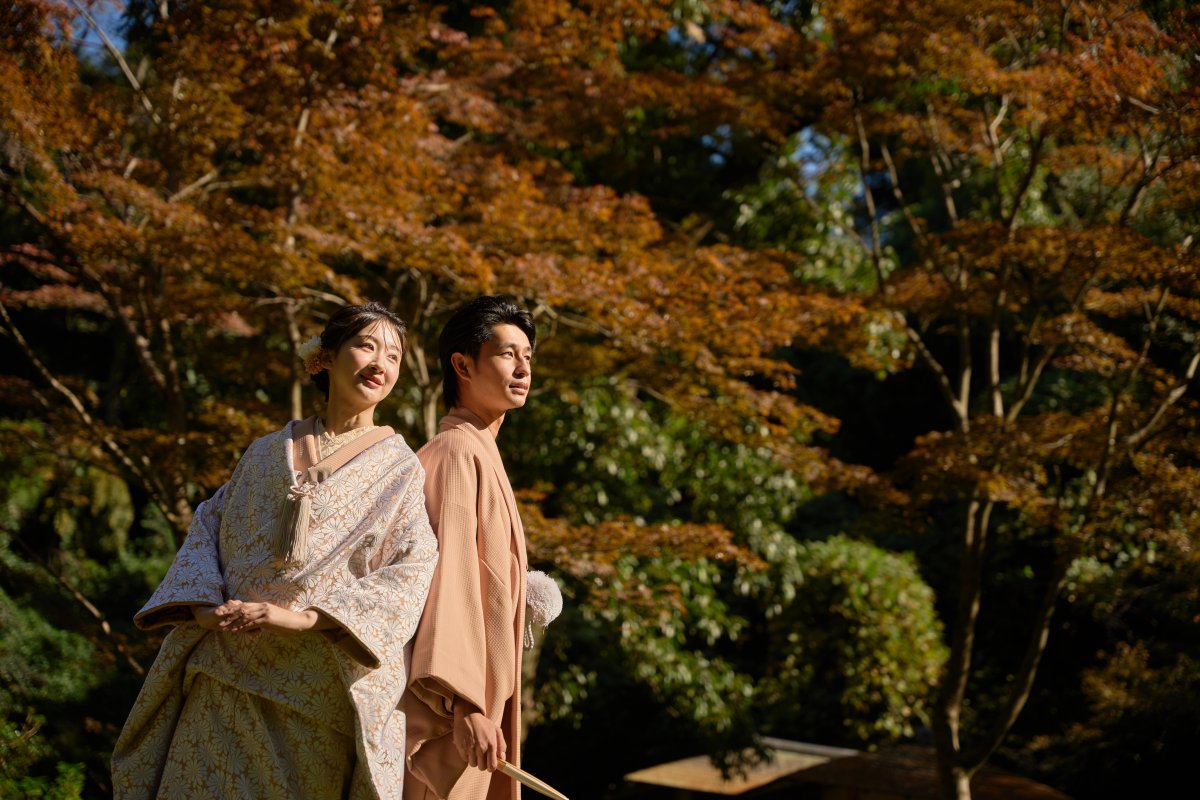 紅葉を背景に佇む淡いピンクの色打掛と羽織袴の新郎新婦