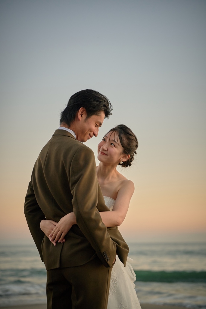 流木に腰掛け夕暮れの海を眺める新郎新婦の後ろ姿