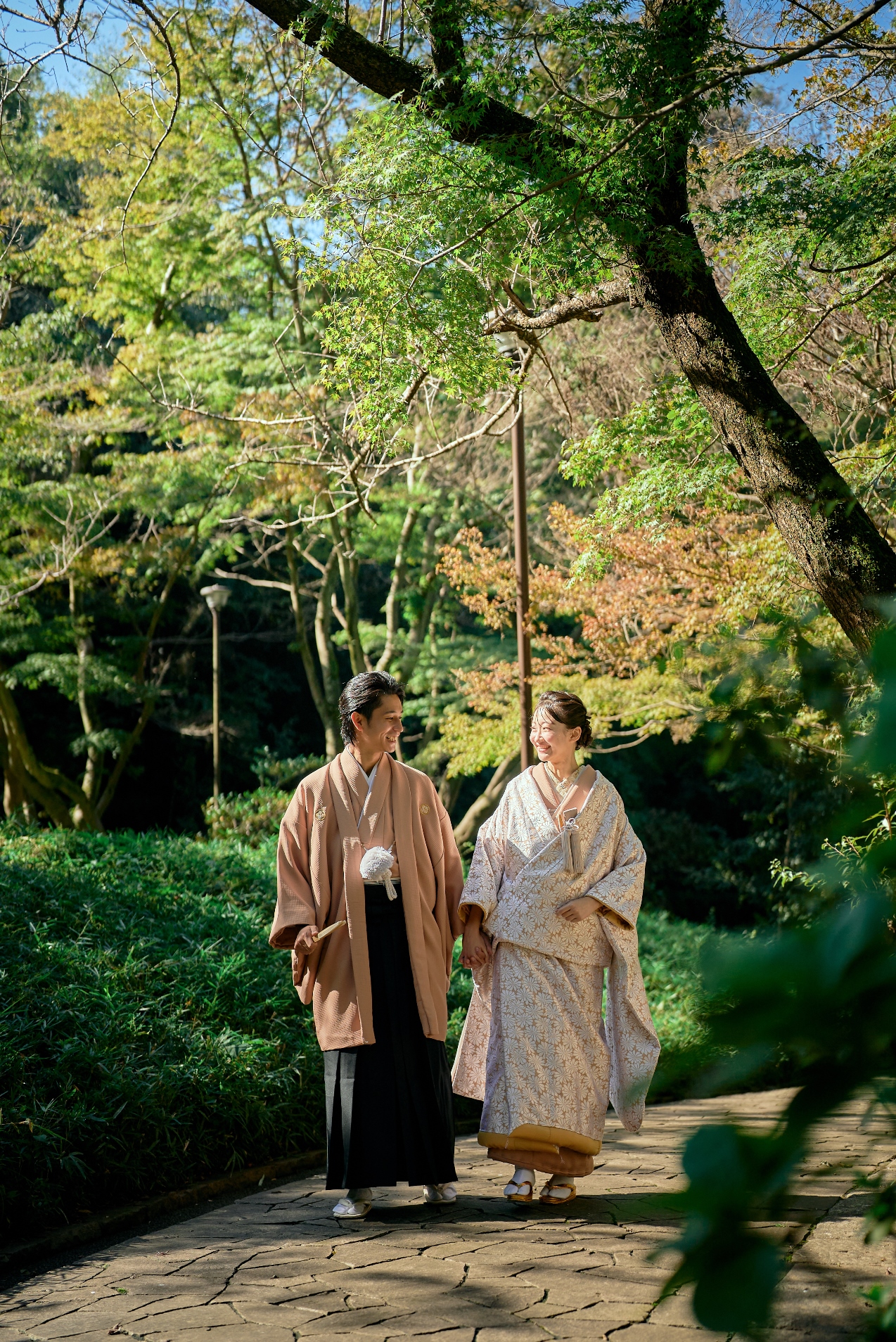 紅葉を背景に佇む淡いピンクの色打掛と羽織袴の新郎新婦