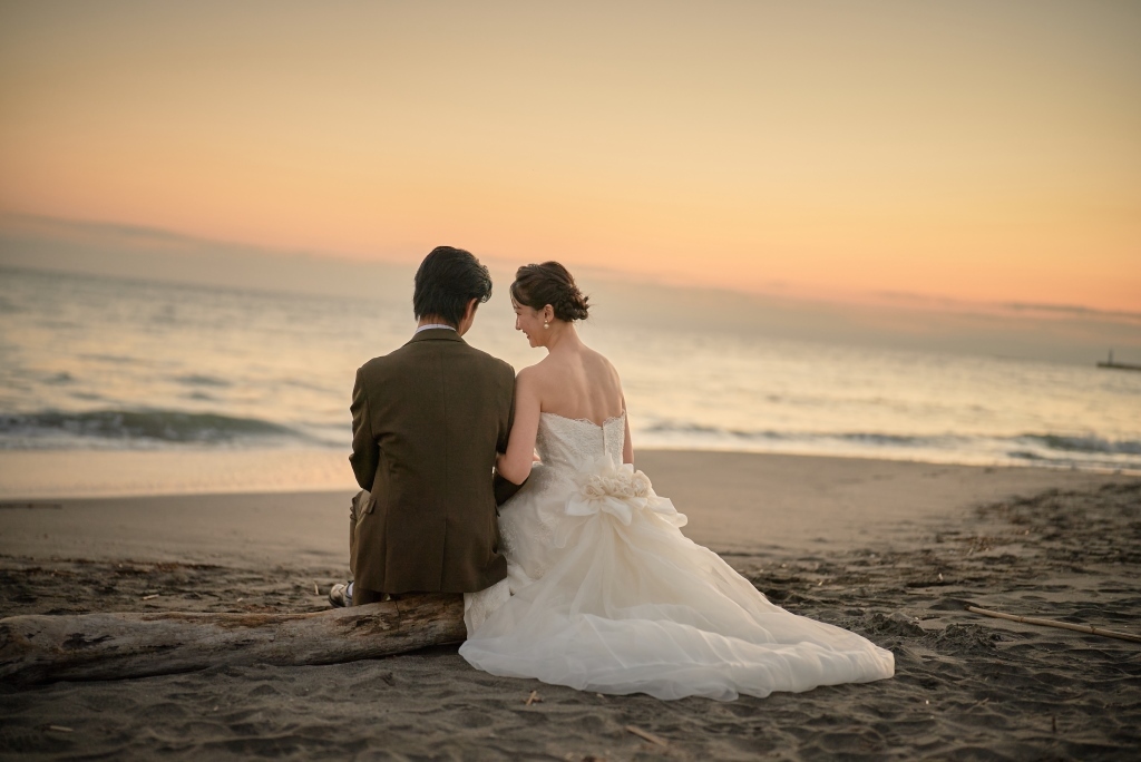 流木に腰掛け夕暮れの海を眺める新郎新婦の後ろ姿