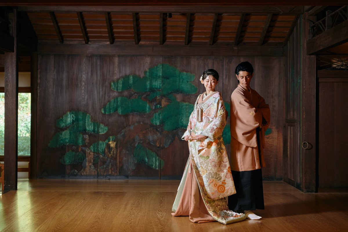 松の絵が描かれた能舞台の壁を背景に背中合わせに立つ新郎新婦
